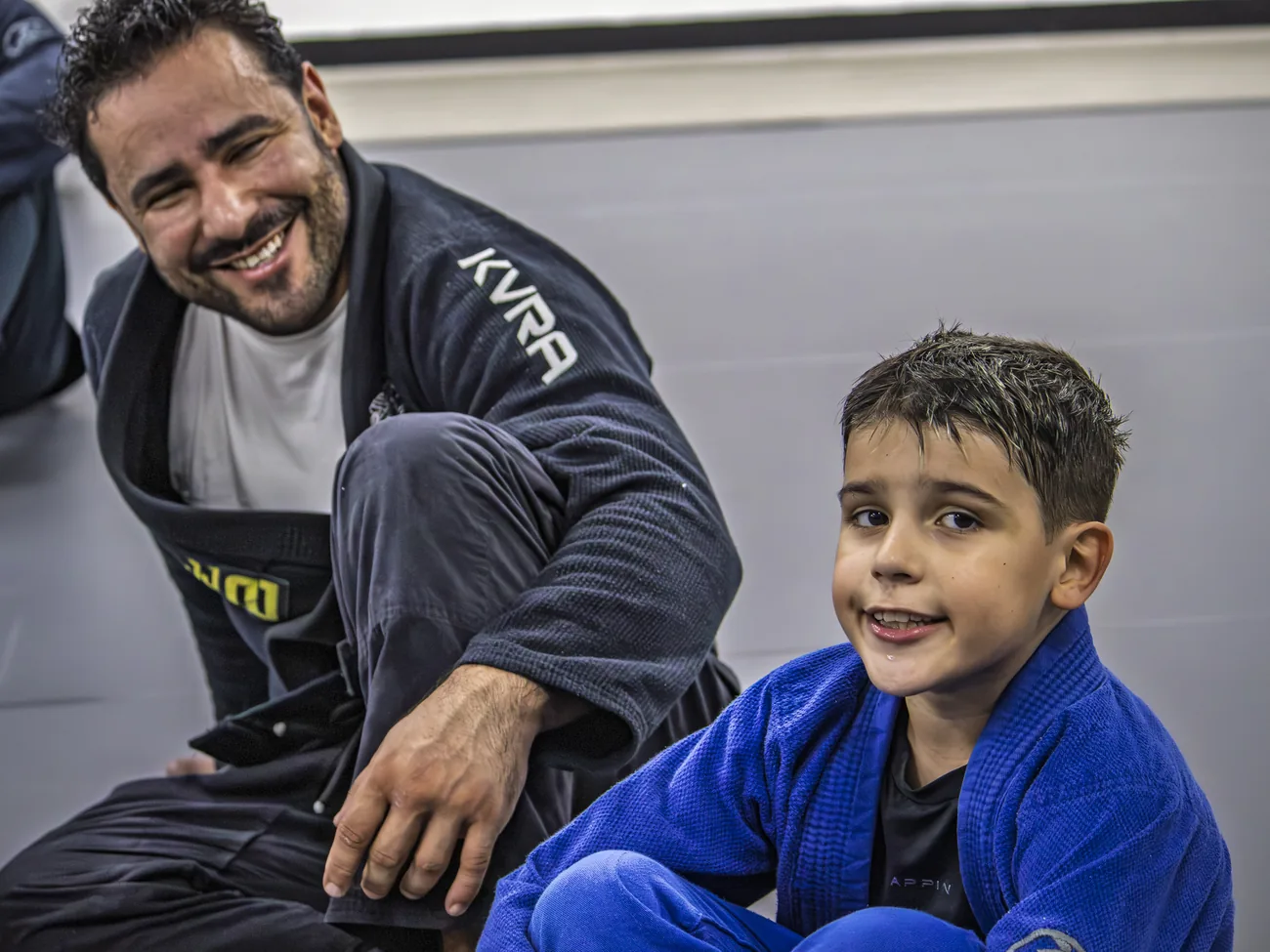 Aluno adulto e criança de kimono sorrindo no tatame